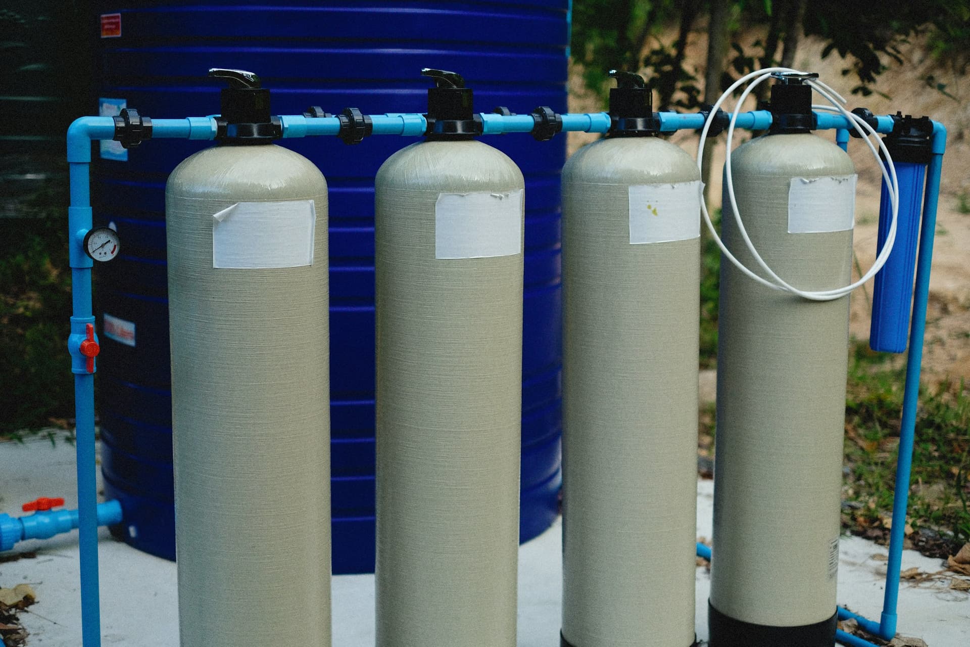 A row of four water filtration tanks with control valves joined by blue pipework, beside a blue cartridge filter housing.